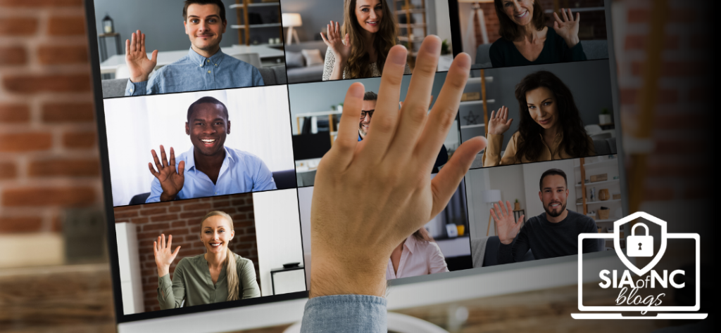 A close-up view of a computer screen displaying a virtual meeting with nine participants, all smiling and waving. A hand from the foreground waves towards the screen, indicating engagement in the virtual interaction.