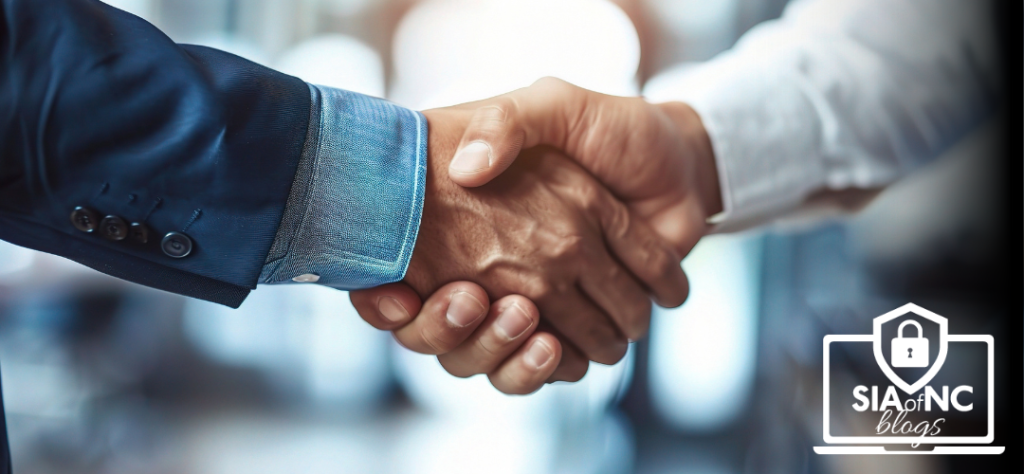 The image shows two people shaking hands in a professional setting, with one wearing a blue suit and the other in a white shirt. The background is blurred, creating a focus on the handshake. A "SIA of NC blogs" logo is placed in the bottom right corner.